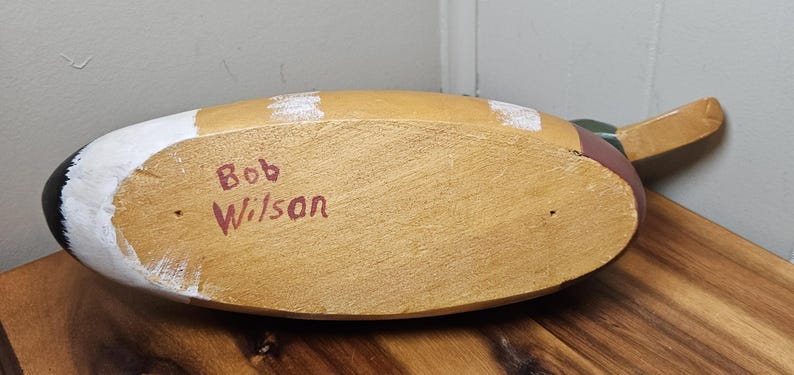 K&ouml;nnte beinhalten: H&ouml;lzerner Entenk&ouml;der mit lackiertem Finish. Die Unterseite ist sichtbar und zeigt den Text "Bob Wilson" in Rot. Die Ente hat einen beigen K&ouml;rper, wei&szlig;e und schwarze Akzente und einen braunen Schwanz.