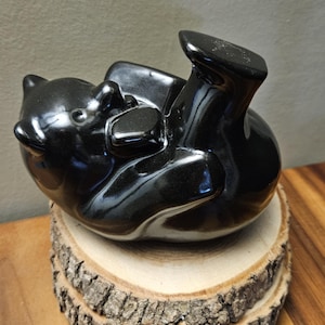 May include: A glossy black bear figurine in a playful pose, lying on its back with its paws in the air. The bear is placed on a natural wood slice, showcasing its smooth, reflective surface. The figurine is a decorative item.