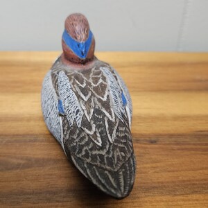 Op de afbeelding: Een gedetailleerd, handgeschilderd eendenbeeldje. De eend heeft een bruine kop met een blauwe streep, een grijs en bruin patroon op het lichaam en blauwe accenten. Het beeldje staat op een houten oppervlak en toont de ingewikkelde details van de vogel.