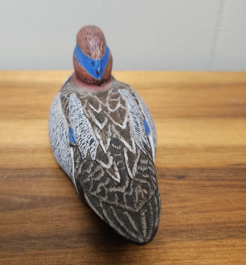 Op de afbeelding: Een gedetailleerd, handgeschilderd eendenbeeldje. De eend heeft een bruine kop met een blauwe streep, een grijs en bruin patroon op het lichaam en blauwe accenten. Het beeldje staat op een houten oppervlak en toont de ingewikkelde details van de vogel.