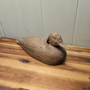 May include: A wooden duck decoy, carved with detailed feathers and a long beak. The duck is a natural brown color and rests on a wooden surface. The decoy is a classic piece of Americana, perfect for rustic decor.