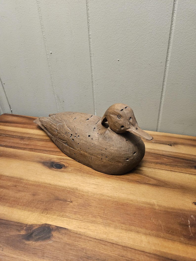 May include: A wooden duck decoy, carved with detailed feathers and a long beak. The duck is a natural brown color and rests on a wooden surface. The decoy is a classic piece of Americana, perfect for rustic decor.