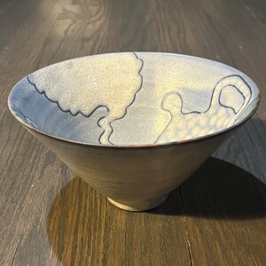 May include: A light blue ceramic bowl with a textured surface and a black design of abstract shapes. The bowl is sitting on a wooden surface.