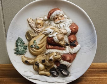 Może przedstawiać: Biały ceramiczny talerz z trójwymiarowym wzorem przedstawiającym Świętego Mikołaja, dziecko, psa i worek z prezentami. Święty Mikołaj ma na sobie czerwony strój z białą brodą i czerwoną czapką. Dziecko ma na sobie różową sukienkę i trzyma białego króliczka. Pies jest brązowo-biały i siedzi obok Świętego Mikołaja. Worek z prezentami jest brązowy i ma zieloną gałązkę ostrokrzewu.
