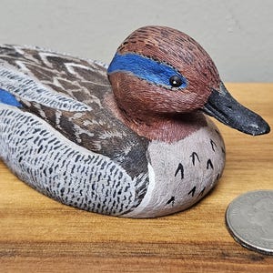 Op de afbeelding: Een handgesneden houten eendenbeeldje met gedetailleerde veermotieven in bruin, grijs en wit. De eend heeft een kastanjebruine kop met een blauwe streep en een zwarte snavel. Er ligt een munt naast de eend.