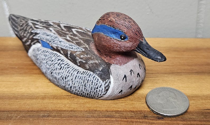 Op de afbeelding: Een handgesneden houten eendenbeeldje met gedetailleerde veermotieven in bruin, grijs en wit. De eend heeft een kastanjebruine kop met een blauwe streep en een zwarte snavel. Er ligt een munt naast de eend.
