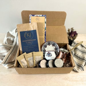 May include: A brown cardboard box filled with a Turkish coffee gift set. The set includes a bag of dark roast coffee, a copper cezve, a small ceramic coffee cup and saucer with a traditional Turkish design, and small jars of spices like cinnamon, ginger, and cloves. The box is filled with brown shredded paper.