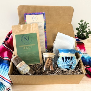 Op de afbeelding: Cadeaudoos met KI Mexico Koffie, een bruine suikerkegel, een blauw-witte keramische mok, kaneelstokjes en theezakjes. De doos bevat ook een kaart met tekst. De items zijn op stro gerangschikt.
