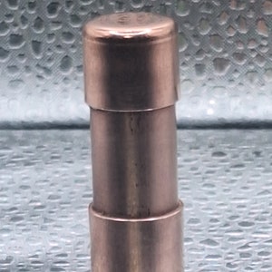 May include: A cylindrical copper-colored object with a three-part design. The top and bottom sections are wider than the central portion. The object has a metallic sheen and sits against a textured, light-colored background.