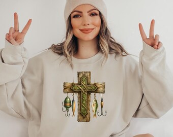 Könnte beinhalten: Beiges Sweatshirt mit einem Design aus einem Holzkreuz, grünen Ranken und Angelködern. Das Kreuz hat ein rustikales, verwittertes Aussehen. Das Sweatshirt ist in einer neutralen Farbe gehalten und hat einen Rundhalsausschnitt.