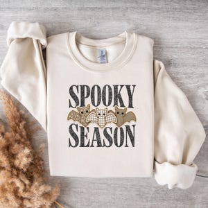 May include: A cream-colored long-sleeve sweatshirt featuring the black text SPOOKY SEASON and a graphic of three bats with patterned wings in shades of brown and beige. The garment has a crew neckline and ribbed cuffs on the long sleeves.