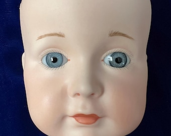 May include: A porcelain doll head with a pale complexion, blue eyes, and delicate painted features. The doll head has subtle blush on the cheeks and a small, closed mouth. The eyebrows are thin and arched, and the head is bald.