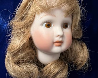 May include: A porcelain doll head with blonde hair, brown eyes, and a painted blush on the cheeks.