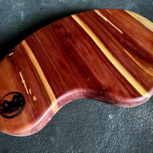 May include: A wooden serving tray with a natural wood grain pattern. The tray is shaped like a crescent moon and has a dark brown stain. The tray has a logo burned into the wood.