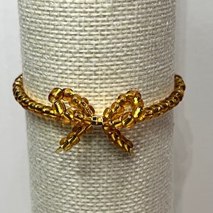May include: A gold beaded bracelet with a bow design. The bracelet is displayed on a white fabric stand.