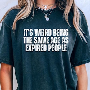 Puede incluir: Camiseta gris oscuro con el texto "IT'S WEIRD BEING THE SAME AGE AS EXPIRED PEOPLE" en blanco. La camiseta es de corte holgado y tiene mangas cortas. La imagen muestra la camiseta en una persona.