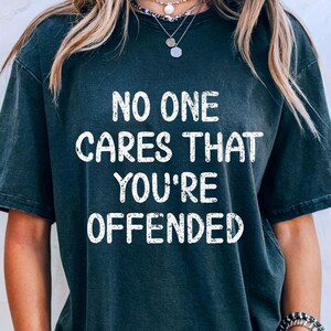 Op de afbeelding: Donkergrijs T-shirt met de witte tekst "NO ONE CARES THAT YOU'RE OFFENDED". Het shirt heeft een licht vervaagde uitstraling. De persoon draagt een spijkerbroekshort en sieraden.