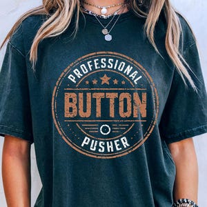 May include: Dark gray t-shirt with a distressed, circular graphic that reads "PROFESSIONAL BUTTON PUSHER" in brown and white. The shirt has short sleeves and a relaxed fit. The model is wearing denim shorts and jewelry.