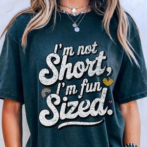 May include: Dark grey t-shirt with the slogan "I'm not Short, I'm fun Sized" in a white, retro font. The shirt has a rainbow and heart graphic. The shirt is worn with denim shorts.