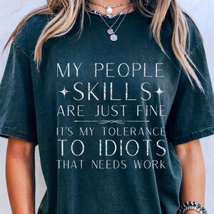 Może przedstawiać: Ciemnoszara koszulka z białym napisem "MY PEOPLE SKILLS ARE JUST FINE IT'S MY TOLERANCE TO IDIOTS THAT NEEDS WORK". Koszulka ma swobodny styl.