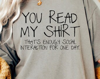 May include: A gray t-shirt with the text "YOU READ MY SHIRT" and "THAT'S ENOUGH SOCIAL INTERACTION FOR ONE DAY" in black. A mustard yellow cardigan is draped over the shoulders. A gold necklace is visible.
