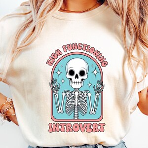 Könnte beinhalten: Cremefarbenes T-Shirt mit Retro-Design und Skelett-Illustration. Das Design enthält die Worte "HIGH FUNCTIONING INTROVERT" in einem rosa und blauen Bogen. Das Shirt wird mit hellblauen Jeans kombiniert.