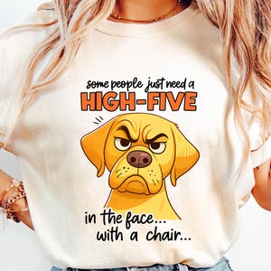 May include: Cream-colored t-shirt featuring a cartoon illustration of a grumpy yellow dog with the text "some people just need a HIGH-FIVE in the face... with a chair...". The shirt is paired with light blue denim jeans.