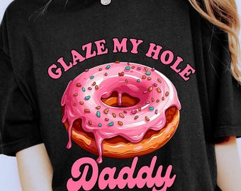 Pode incluir: Camiseta preta com um desenho de donut rosa glaceado e granulado. O texto "GLAZE MY HOLE Daddy" é rosa. A camiseta é usada com shorts com estampa de leopardo.