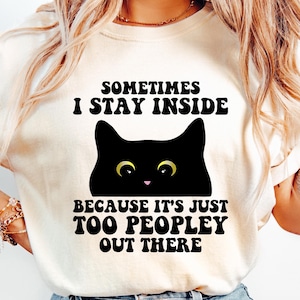 May include: A cream-coloured t-shirt featuring the text "SOMETIMES I STAY INSIDE BECAUSE IT'S JUST TOO PEOPLEY OUT THERE" and a black cat graphic with yellow eyes. The shirt is worn with blue jeans.