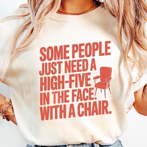 May include: Cream-colored t-shirt with the text "SOME PEOPLE JUST NEED A HIGH-FIVE IN THE FACE WITH A CHAIR" in red. The design includes a red chair graphic. The shirt is paired with light blue jeans.