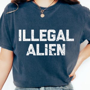 May include: A navy blue t-shirt with the words "ILLEGAL ALIEN" printed in distressed white lettering. The shirt is paired with light wash denim jeans and white sneakers. The overall style is casual and relaxed.