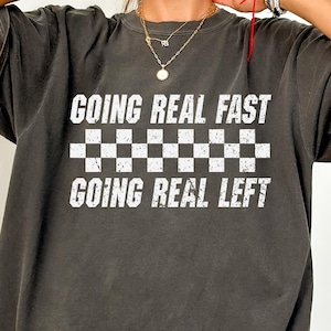 Può includere: T-shirt grigio scuro con la scritta "GOING REAL FAST" sopra un motivo a scacchi bianchi e neri, e "GOING REAL LEFT" sotto. La maglietta ha maniche corte e una vestibilità ampia.