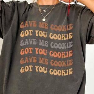 Può includere: T-shirt grigio scuro con la scritta "GAVE ME COOKIE GOT YOU COOKIE" in un design retrò a strati. Il testo è in tonalità di marrone, arancione e grigio. La maglietta ha maniche corte e una vestibilità ampia.