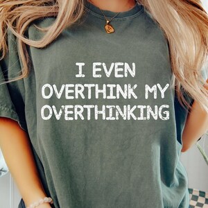 May include: Dark green t-shirt with the text "I EVEN OVERTHINK MY OVERTHINKING" in white. The shirt has a relaxed fit and short sleeves. The person is wearing dark grey leggings and a bracelet.