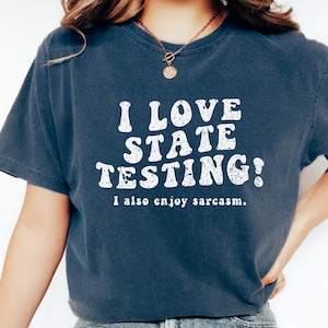 May include: Dark blue t-shirt with the text "I LOVE STATE TESTING! I also enjoy sarcasm." The shirt is paired with light wash denim jeans and white sneakers. The shirt's text is in a bold, white font.
