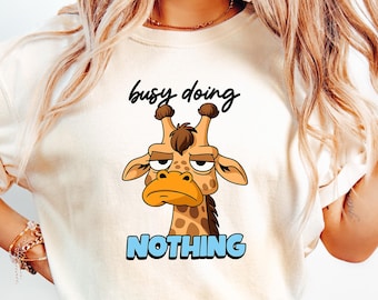 Może przedstawiać: Kremowy t-shirt z kreskówkową żyrafą z ponurym wyrazem twarzy. Tekst "busy doing" znajduje się nad żyrafą, a "NOTHING" pod nią w niebieskich literach. Koszulka jest połączona z jasnoniebieskimi dżinsami.