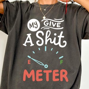 Könnte beinhalten: Dunkelgraues T-Shirt mit dem Text "MY GIVE A SHIT METER" in Weiß und Rot. Das Design beinhaltet eine Tankanzeige, deren Nadel auf leer zeigt. Das Shirt hat kurze Ärmel und eine lockere Passform.