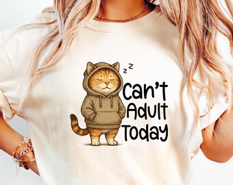 May include: Cream-colored t-shirt featuring a cartoon cat wearing a brown hoodie. The cat has closed eyes and the text "Can't Adult Today" in black. The shirt is paired with light blue denim jeans.