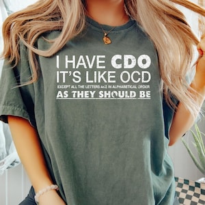 May include: Dark green t-shirt with the text "I HAVE CDO IT'S LIKE OCD EXCEPT ALL THE LETTERS ARE IN ALPHABETICAL ORDER AS THEY SHOULD BE" in white. The shirt is a casual style.