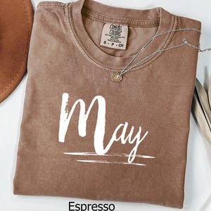 May include: Espresso-colored t-shirt with the word "May" in white script. The shirt has a crew neck and a small tag at the neckline. A silver necklace with a small pendant is draped across the shirt. A dried plant and a brown hat are in the background.
