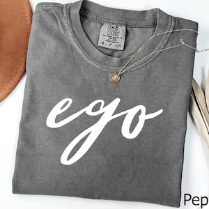 Puede incluir: Una camiseta color pimienta con la palabra "ego" en escritura blanca. Un collar dorado con un pequeño colgante descansa sobre la camiseta. La camiseta tiene cuello redondo y una etiqueta "Comfort Colors".