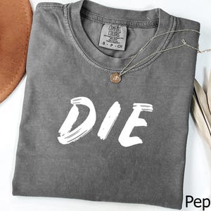May include: Pepper-colored t-shirt with the word "DIE" in white brushstroke lettering. The shirt has a crew neck and short sleeves. A gold-colored necklace with a pendant is draped across the shirt.