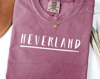 May include: Berry-colored t-shirt with the word "NEVERLAND" in white block letters. A gold necklace with a pendant rests on the shirt. The shirt has a crew neck and a small tag at the neckline.