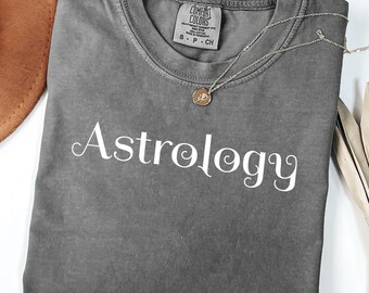 Könnte beinhalten: Ein graues T-Shirt mit dem Wort "Astrology" in weißer Schrift. Eine goldene Anhänger-Halskette liegt auf dem Shirt. Das Shirt ist gefaltet, und ein brauner Hut ist im Hintergrund zu sehen. Das Shirt ist mit "Pepper" beschriftet.