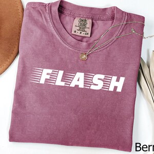 Puede incluir: Camiseta de color bayas con la palabra "FLASH" en blanco, estilizada con líneas de velocidad. La camiseta tiene cuello redondo y una pequeña etiqueta en el cuello. Un collar dorado con un colgante está drapeado sobre la camiseta. La palabra "Berry" está en la esquina inferior derecha.
