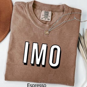 May include: Espresso-colored t-shirt with the word "IMO" in bold white letters with a black shadow. The shirt has a crew neck and a small tag. A silver necklace with a pendant is draped across the shirt.