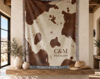 May include: A woven throw blanket featuring a brown and cream cowhide pattern with fringed edges. The blanket is personalized with the letters C&M and the date JULY 24, 2027, printed in a dark brown serif font on the lower right side.