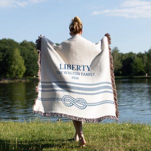 May include: A cream-coloured throw blanket with navy blue nautical rope and stripe design. The blanket has the word "LIBERTY" and "THE WALTON FAMILY 2026" printed in navy blue. The blanket has a brown fringe.