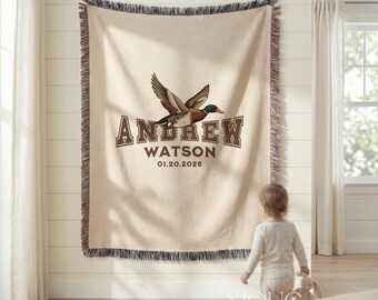 May include: A beige woven throw blanket featuring a central illustration of a flying mallard duck. Below the duck, the text reads ANDREW WATSON and the date 01.20.2026 in brown block lettering. The blanket has a fringed edge on the top and bottom.