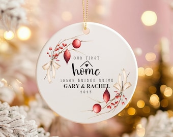 May include: A white ceramic Christmas ornament with the text "OUR FIRST home" and address details. The design includes red berries, leaves, and a bow. A gold string is attached for hanging.